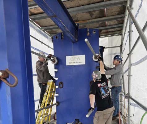 Workers installing or servicing a large industrial heat exchanger unit, using tools and ladders, with safety gear on.